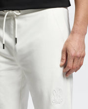 A man wears Psycho Bunny MENS LEVANZO EMBROIDERED MICRO FRENCH TERRY SWEATSHORTS (B6R578F200) in soft white micro French terry, with a drawstring waist and embroidered bunny and crossbones logo on the upper thigh. Shown mid-torso to hand.