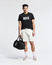 A young man stands against a plain background, wearing a black graphic tee and Psycho Bunny MENS LEVANZO EMBROIDERED MICRO FRENCH TERRY SWEATSHORT (B6R578F200), paired with white crew socks, beige sneakers, and holding a black duffel bag in his left hand.
