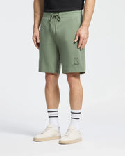 A person wears Psycho Bunny's Levanzo Embroidered Micro French Terry Sweatshorts with a bunny logo, paired with a black shirt, white socks with black stripes and text, and light beige sneakers against a light gray backdrop.