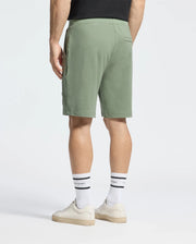 A person is seen from the back wearing Psycho Bunny's olive green Mens Levanzo Embroidered Micro French Terry Sweatshort, a black shirt, and white socks with black stripes. White sneakers enhance the look against a plain white background, subtly highlighted by an embroidered bunny.