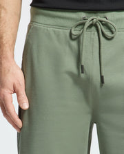 A person wears Psycho Bunny's Men's Levanzo Embroidered Sweatshorts in sage green, made from micro French terry with a drawstring waist. Their hand beside them subtly reveals a black shirt, exuding effortless comfort.