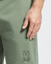 A close-up shows a hand resting on green sweatpants made from micro French terry. The Psycho Bunny's Mens Levanzo Sweatshort, B6R578F200, features an embroidered bunny head above crossed bones near the pocket, set against a plain white background.