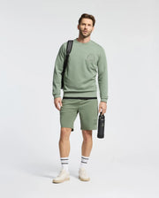A man wearing Psycho Bunny's Men's Levanzo Embroidered Micro French Terry Sweatshort, a green sweatshirt, holds a water bottle and sports white sneakers with striped socks. He also carries a black backpack while standing against a plain background, exuding comfort and style.