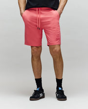 A person stands with hands in pockets, wearing Psycho Bunny MENS LEVANZO EMBROIDERED MICRO FRENCH TERRY SWEATSHORT - B6R578F200 in salmon pink, a black shirt, black socks, and black Adidas sneakers against a plain light gray background.