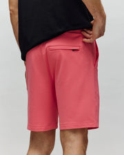 A person in a black shirt and Psycho Bunny MENS LEVANZO EMBROIDERED MICRO FRENCH TERRY SWEATSHORT - B6R578F200 stands with one hand on their hip, showing off the bright pink shorts’ micro French terry fabric and zippered back pocket.
