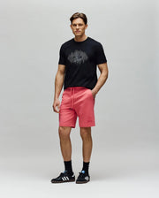 A man with short brown hair and a neutral expression wears a black graphic tee, black socks, black Adidas sneakers, and bright pink Psycho Bunny MENS LEVANZO EMBROIDERED MICRO FRENCH TERRY SWEATSHORT - B6R578F200.