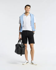 A person poses against a plain background, donning a white polo, versatile shorts, and white sneakers. Draped over their shoulders is Psycho Bunny's light blue MENS SHIRO KNIT HONEYCOMB SHORT. They hold a black bag in one hand, effortlessly transitioning from casual to chic.