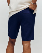 A person, seen from behind with a hand on their hip, wears a cream shirt and Psycho Bunny MENS SHIRO KNIT HONEYCOMB SHORT - B6R585F200 in navy blue. The shorts have a honeycomb piqué texture and a single back pocket.