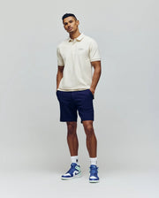 A man poses confidently against a light gray background wearing a cream polo, Psycho Bunny MENS SHIRO KNIT HONEYCOMB SHORTS, white socks, and blue-and-white sneakers. An embroidered Bunny logo adds a playful touch to his look.