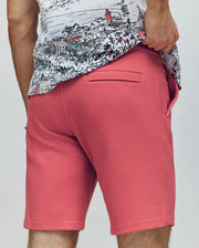 A person, back to the camera, wears Psycho Bunny's coral pink MENS SHIRO KNIT HONEYCOMB SHORT (B6R585F200) and a white honeycomb-patterned piqué shirt, with their right hand in their pocket and leg hair visible.