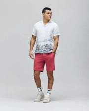 A man stands against a plain background in a white short-sleeve shirt with an embroidered Bunny cityscape, Psycho Bunny MENS SHIRO KNIT HONEYCOMB SHORT - B6R585F200 in pink, white socks with black stripes, and white sneakers.