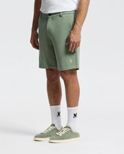 A person wearing Psycho Bunny's Shiro Knit Honeycomb Shorts, B6R585F200, and matching sneakers stands against a plain background. The light green shorts, made from honeycomb-patterned piqué, have side pockets and an embroidered detail near the hem, making them a versatile wardrobe addition.