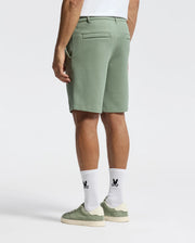 A person in Psycho Bunny's MENS SHIRO KNIT HONEYCOMB SHORT (B6R585F200) in olive stands against a white background, paired with white socks featuring a black logo and green-and-beige sneakers, styled with hands by their sides and shown from the back.