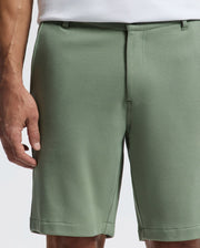 A person is shown from the waist down wearing Psycho Bunny's MENS SHIRO KNIT HONEYCOMB SHORT - B6R585F200 in light green piqué. They are also in a white shirt, with their left arm partially visible against a plain white background.