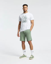 A man poses against a plain, light background in a white graphic T-shirt, versatile green MENS SHIRO KNIT HONEYCOMB SHORT by Psycho Bunny, matching green sneakers, and white socks with black details.