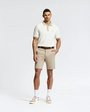 A man stands against a white background wearing Psycho Bunny's MENS SHIRO KNIT HONEYCOMB SHORT (B6R585F200) with a brown belt, white crew socks, and beige sneakers. One hand is in his pocket as he faces forward with a neutral expression.