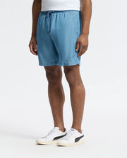 A person models the MENS KERVIN LIGHTWEIGHT DENIM WOVEN SHORT - B6R633F200 by Psycho Bunny, featuring blue drawstring shorts with an elasticized waist and white sneakers with black accents, against a plain background.