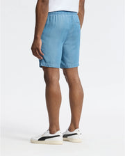 A person wearing Psycho Bunny's MENS KERVIN LIGHTWEIGHT DENIM WOVEN SHORT in light blue and white sneakers with black detailing stands with their back to a plain white background.