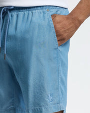 A person wears Psycho Bunny's light blue MENS KERVIN LIGHTWEIGHT DENIM WOVEN SHORTS—featuring an elasticized waist, drawstring, and a subtle embroidered Bunny logo—and pairs them with a white top against a plain white background, with their hand resting in the pocket.