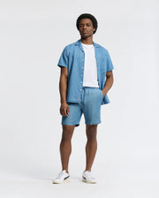 A man with short curly hair wears a Psycho Bunny Kervin lightweight denim short-sleeve shirt and matching shorts with an elastic waist over a white T-shirt, completed by white sneakers. An embroidered bunny peeks from his pocket as he stands against a plain background.
