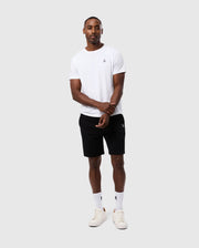 A man stands against a plain background wearing a white t-shirt, Psycho Bunny's regular fit MENS LEVY FLEECE SWEATSHORT (B6R862D200) in black, white socks, and white sneakers. He has short hair and is posing with his hands clasped together.