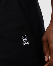 Close-up of a person's hand beside the pocket of Psycho Bunny's MENS LEVY FLEECE SWEATSHORT - B6R862D200, featuring an embroidered logo of a cartoon skull and crossbones with bunny ears. The background is light gray.