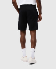 Rear view of a person wearing a white T-shirt, MENS LEVY FLEECE SWEATSHORT - B6R862D200 by Psycho Bunny in black cotton, white socks, and white sneakers. The person is standing on a light gray background with their hands by their sides.