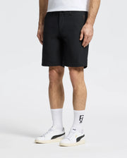 A person in breathable Psycho Bunny MENS 10K STEP SHORT - B6R969F200 black shorts, a white T-shirt, and white socks with black designs, stands in white sneakers on a plain white background.