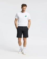 A man wearing a Psycho Bunny white t-shirt with a small green graphic and breathable MENS 10K STEP SHORT black shorts (B6R969F200) stands with hands in pockets. He's also in white socks and sneakers against a plain white background.