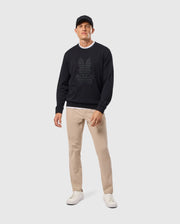 A man wearing a Psycho Bunny MENS KINGSTON SWEATSHIRT - B6S480D200 in black, matched with beige pants and white sneakers, stands against a light background. He looks directly ahead with a neutral expression.