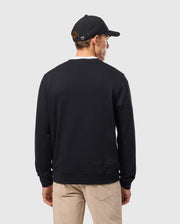 A person wearing a Psycho Bunny MENS KINGSTON SWEATSHIRT - B6S480D200 in black, paired with a black baseball cap and beige pants, stands with their back to the camera against a plain gray background.