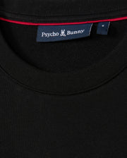 Close-up of a black cotton jersey shirt showcasing the "Psycho Bunny" brand label with a small bunny logo. The label is navy with white text, and there is a red accent inside the collar. The product is identified as the MENS KINGSTON SWEATSHIRT - B6S480D200 by Psycho Bunny.