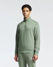 A man in the Psycho Bunny MENS DESERT 1/4 MOCK ZIP - B6S488F200, a light green 100% cotton quarter-zip pullover with matching pants, both with an embroidered wordmark logo on the chest and thigh, stands against a plain white background.
