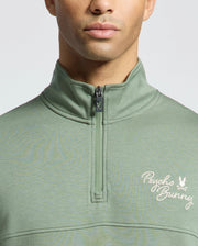 A man is wearing the Psycho Bunny Men's Desert 1/4 Mock Zip (B6S488F200) in light green, featuring "Psycho Bunny" and an embroidered bunny logo on the left chest. The plain, light background accentuates its 100% cotton fabric for ultimate comfort.