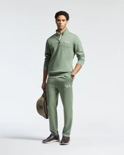 A man wears the Psycho Bunny MENS DESERT 1/4 MOCK ZIP tracksuit, paired with matching pants featuring an embroidered wordmark logo. He holds a brown helmet under his arm, dons dark brown shoes, and stands against a plain light background.