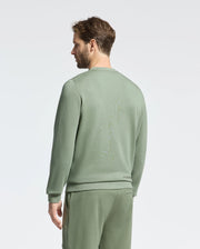 A man stands against a plain backdrop in a sage green Psycho Bunny Melbourne Embroidered Classic Modal Crewneck Sweatshirt, paired with matching pants. Facing sideways, he looks left with short hair and a beard, embodying relaxed elegance.