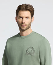 A man with short brown hair and a beard is wearing the Psycho Bunny Men's Melbourne Embroidered Classic Modal Crewneck Sweatshirt, B6S561F200, in light green. It features an embroidered cartoon bunny patch and is made of a breathable Modal blend for comfort on its plain backdrop.
