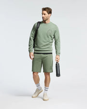 A man in a Psycho Bunny light green Melbourne Embroidered Classic Modal Crewneck sweatshirt and matching shorts holds a black water bottle and bag. He's wearing white socks with black stripes and beige sneakers against a plain white background.