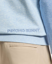Close-up of a person wearing the Psycho Bunny MENS ARCHIE MELANGE COLOR BLOCK SWEATSHIRT, B6S630F200, in light blue with an embroidered Bunny logo at the hem, paired seamlessly with beige pants for a subtle monochrome styling.