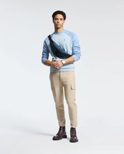 A man poses against a plain backdrop in monochrome style, wearing a light blue Psycho Bunny ARCHIE melange color block sweatshirt, beige cargo pants, and brown boots. His black crossbody bag features an embroidered Bunny, and he wears a watch on his left wrist. He gazes ahead with ease.
