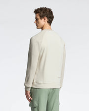 A person in a Psycho Bunny MENS ARCHIE MELANGE COLOR BLOCK SWEATSHIRT - B6S630F200 stands sideways, wearing green cargo pants and gazing down. The simple studio backdrop complements the outfit's understated elegance.