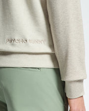A person wears the ARCHIE MELANGE COLOR BLOCK SWEATSHIRT by Psycho Bunny, featuring a beige cotton design with "PSYCHO BUNNY" and an embroidered bunny. The sleeve complements their light green pants against a plain backdrop.