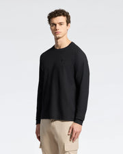 A person with curly hair stands against a plain white background, wearing a Psycho Bunny MENS TAYLOR PIQUE LONG SLEEVE TEE and beige cargo pants. Their neutral expression seamlessly combines casual comfort with understated style.