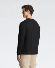 A person with curly hair stands facing away, wearing a Psycho Bunny MENS TAYLOR PIQUE LONG SLEEVE TEE (B6T580F200) in black and beige cargo pants against a plain, light gray background.