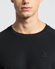 The MENS TAYLOR PIQUE LONG SLEEVE TEE by Psycho Bunny is a sleek black sweater crafted from soft Pima cotton. It features an embroidered rabbit and crossbones logo on the chest, set against a plain white background to highlight its luxurious texture.