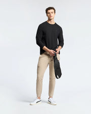 A man stands against a plain background wearing a Psycho Bunny MENS TAYLOR PIQUE LONG SLEEVE TEE in black, beige cargo pants, and white sneakers. He holds a black crossbody bag in front of him.