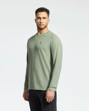 A man with short curly hair, dressed in a sage green Psycho Bunny MENS TAYLOR PIQUE LONG SLEEVE TEE and black pants, stands against a light gray background, looking directly forward.