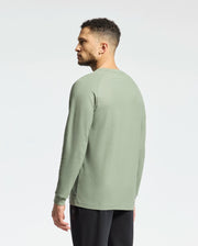 A man with short curly hair wears a light green Psycho Bunny Taylor Pique long-sleeve tee (B6T580F200) and black pants, facing slightly away from the camera against a plain white background.