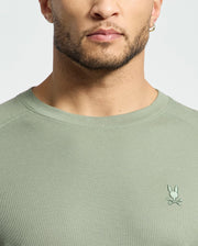 Close-up of a person wearing the Psycho Bunny MENS TAYLOR PIQUE LONG SLEEVE TEE - B6T580F200 in light green. The soft Pima cotton tee features an embroidered Bunny logo over crossbones on the chest, focusing on the torso with a partially visible face.