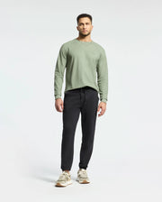 A person wears a Psycho Bunny MENS TAYLOR PIQUE LONG SLEEVE TEE in light green, paired with black jogger pants and beige and white sneakers. An embroidered Bunny adds playfulness to the look against a plain white background.